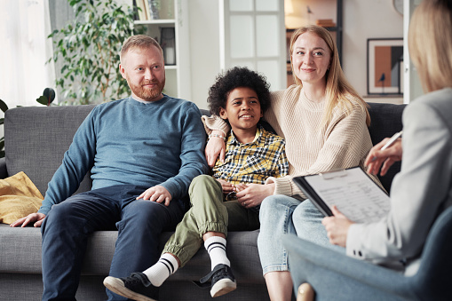 Young parents with adoptive child sitting on sofa and talking to social worker during their meeting at home