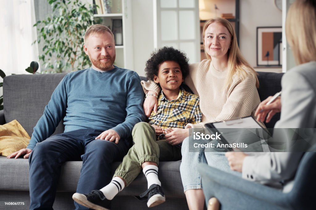 Adoptive family meeting with social worker Young parents with adoptive child sitting on sofa and talking to social worker during their meeting at home Foster Care Stock Photo