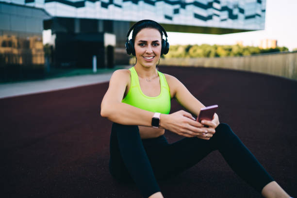 Portrait of cheerful caucasian female athlete in headphones looking at camera using gadgets for entertainment on training, 20s happy sporty girl listen music via smartphone and sound accessory retrato de una alegre atleta caucásica con auriculares mirando a la cámara usando dispositivos para el entretenimiento en el entrenamiento, chica deportiva feliz de los años 20 escucha música a través de un teléfono inteligente y un accesorio de soni
