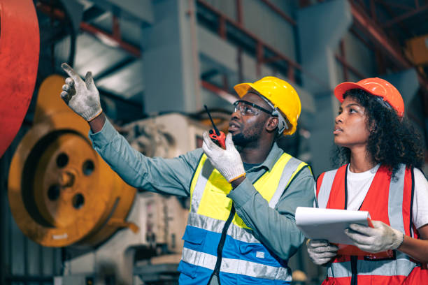 istock Two professional engineer/worker/technician use clipboard discuss work, walk in steel metal manufacture factory plant industry. Black African American man and woman wear hard hat check quality machine 1408062279