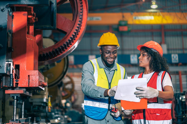 istock Two professional engineer/worker/technician use clipboard discuss work, walk in steel metal manufacture factory plant industry. Black African American man and woman wear hard hat check quality machine 1408062307