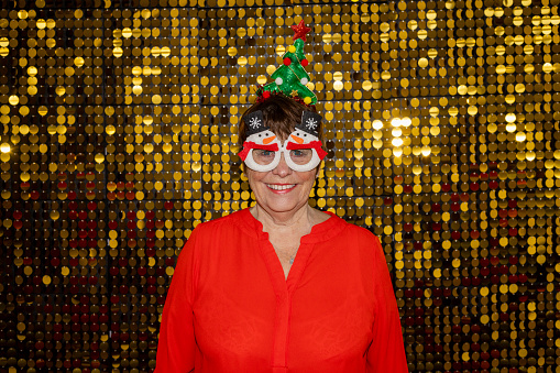Christmas party-themed portraits on gold shiny sequence background shot in a studio. Senior woman looking at camera smiling wearing novelty Christmas glasses and hat.