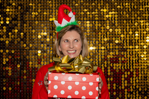 Christmas party-themed portraits on gold shiny sequence background shot in a studio. Woman holding a gift box and wearing a Christmas novelty headband.