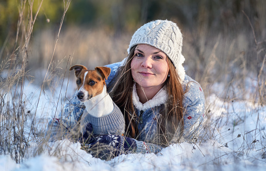 Young woman in winter jacket lying down at snow covered ground, holding her Jack Russell terrier dog, blurred trees background