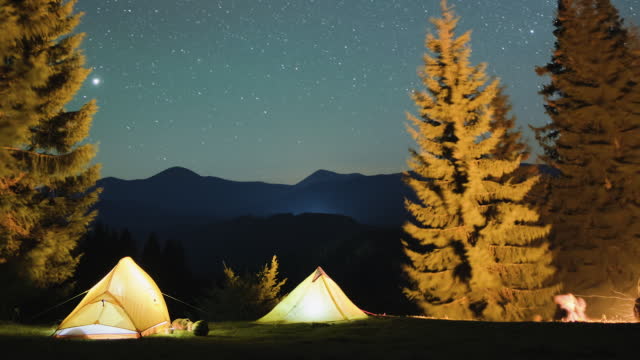 Timelapse of hikers resting besides bright bonfire near illuminated tourist tents on camping site in mountain woods under night sky with sparkling stars. Active lifestyle and outdoor living concept