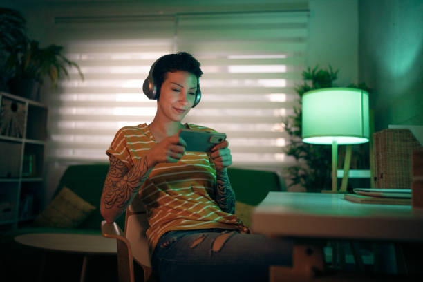 современная кавказская молодая женщина играет в видеоигры на мобильном телефоне - headphones women tattoo music стоковые фото и изображения