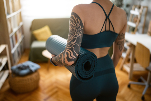 Rear-view of unrecognizable Caucasian young woman with tattoos, holding yoga mat, ready for home workout