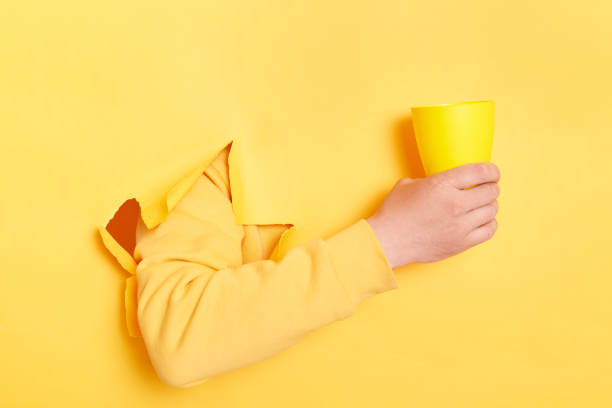Human's holding in hand cup of coffee, tea or water isolated through torn yellow wall. Copy space advertisement place for text image promotional content Advertising area workspace mock up
