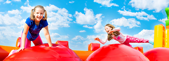 Two happy little girls having lots of fun while jumping from ball to ball on an inflate castle during summer sunny day. Panoramic.