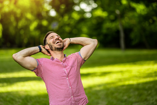 Cheerful man standing with arms behind head while relaxing in nature on beautiful sunny summer day. Copy space.