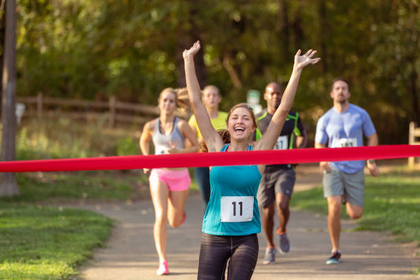 A young adult female athlete celebrates with arms raised when crossing the finish line in first place when running in a race with other men and women on a warm and sunny day.