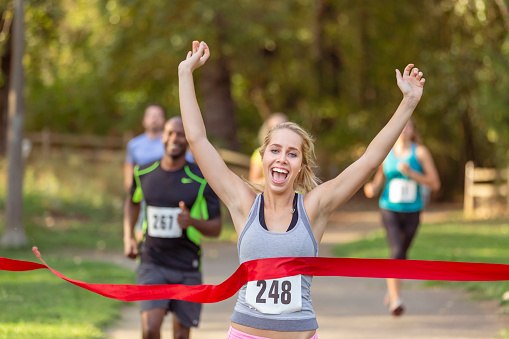 A young adult female athlete celebrates with arms raised when crossing the finish line in first place when running in a race with other men and women on a warm and sunny day.