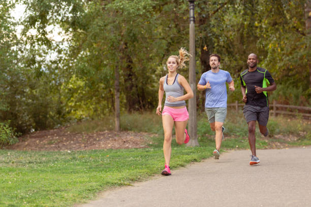 A group of men and women who are in a running club enjoy running together along a paved path through a forested park on a warm and sunny day.