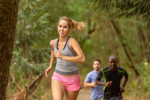 A group of men and women who are members of a running club enjoy a trail run along a dirt path through a forest together. Selective focus on a young woman at the front of the group.
