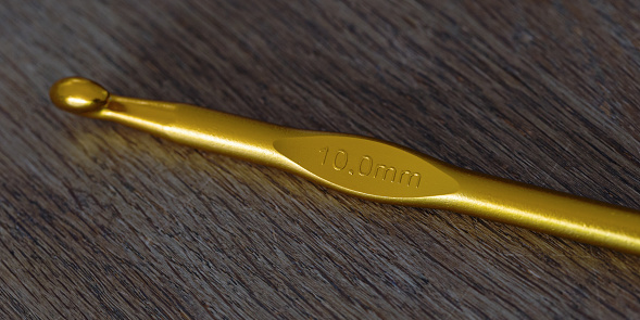 Hihg angle macro close-up of a gold colored plastic 10 mm crochet hook laying on a wooden table