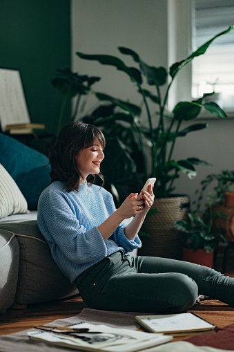 Smiling woman with dental braces typing text message on her smartphone while sitting on the floor in the living room.