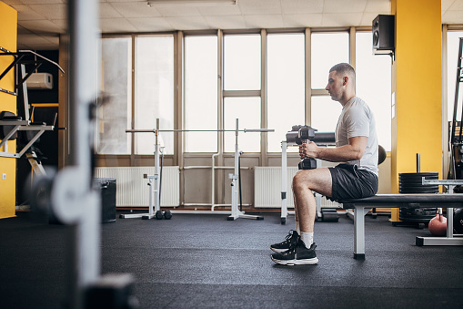 One man, fit man sitting on weight bench and training with dumbbells in gym alone.