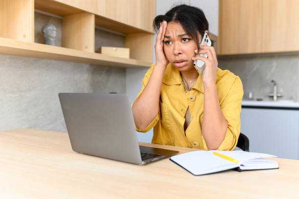 istock Annoyed young woman talks on the smartphone using laptop computer for remote work from home, irritated female looks at the laptop screen 1408119659