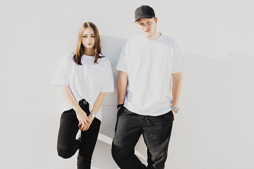 Young fashionable couple wearing designer t-shirts (property released) and black trousers standing outdoors on concrete wall. Looking Towards the Camera. Millennial Generation Male - Female Young Urban Fashion Vintage Portrait.