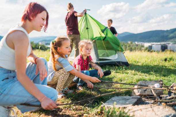 istock Three sisters smiling and roasting marshmallows and candies on sticks over a campfire flame while two brothers set up the green tent. Happy family outdoor picnic camping activities concept. 1408127830