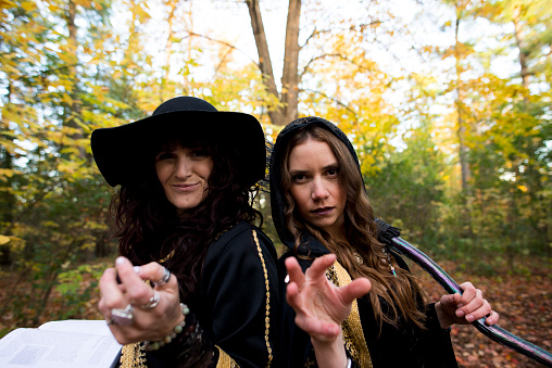 Two young woman wearing fashionable witch costumes and holding a spell book and broomstick are holding out their hands menacingly towards the camera as if casting a magic spell on Halloween.