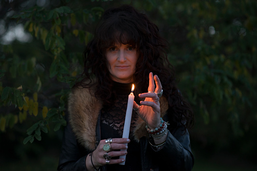 Young woman dressed up in witch robe costume is holding her hand out as if enchanting the burning tall white candle she is holding in her other hand in the middle of a dark forest on Halloween. She is staring in a creepy way directly at the camera.
