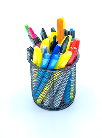 Many colored ink pens and highlighters are stacked in a metal desktop pen holder on a white tabletop studio set, representing business and art.