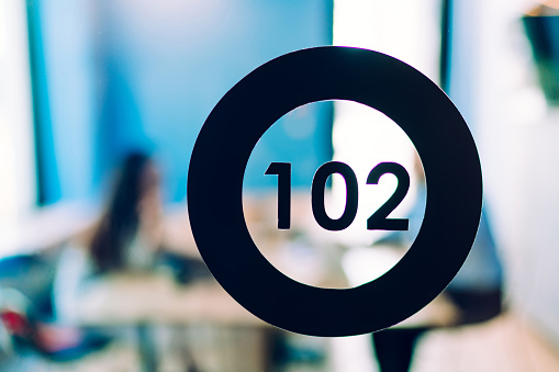 Closeup of black circle with number 102 inside indicated on glass wall of office room with people sitting at table on background