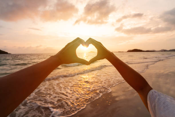 日没時にビーチで手でハートの形を作る若いカップルの旅行者、新婚旅行の恋人、バレンタインデーのコンセプト - ハッピー ストックフォトと画像