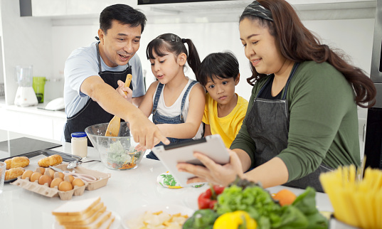 Happy asian parents and their kids having fun while cooking and following recipe on the internet.