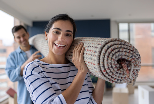 Portrait of a happy Latin American couple moving house and carrying a rolled up carpet - lifestyle concepts