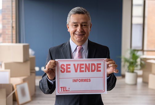 Portrait of a happy Latin American Real Estate Agent holding a for sale sign in Spanish at an apartment - real estate agent concepts