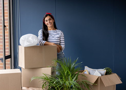 Happy Latin American woman moving house and packing in boxes while looking at the camera smiling