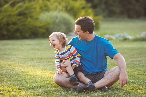 Father and son playing and laughing together, sitting on grass in green park on summer day. Happy family concept.
