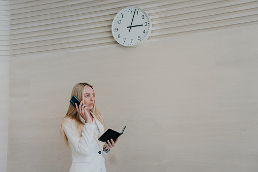 Worried blonde businesswoman at office talking by phone with diary standing under wall clock. Italian beautiful manager negotiates with supplier. Unhappy boss asking by phone employee for being late.