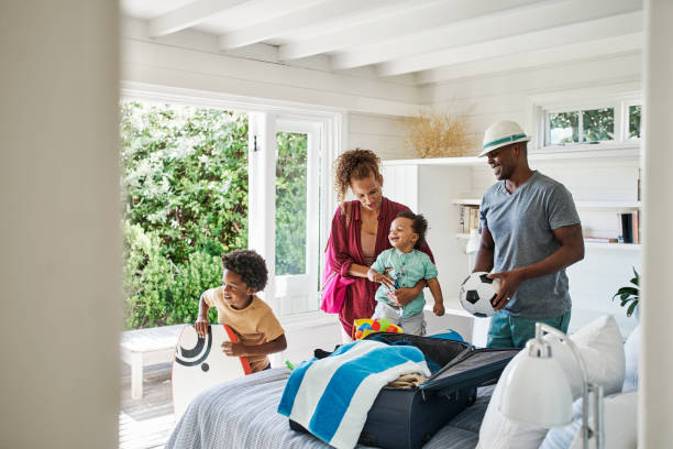 família negra alegre fazendo as malas para férias em família em sua casa ou um quarto de hotel. pais e dois filhos colocando toalhas e brinquedos em uma mala para passar férias na praia. hora de viajar e se divertir - beach suitcase vacations summer - fotografias e filmes do acervo