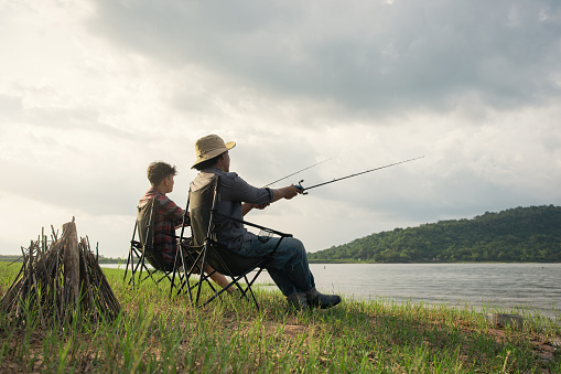 Angler friendship sitting a chair and fishing together at the lake. Camping fire on the shore of the lake. Fisherman with bonfire. Survival concept.