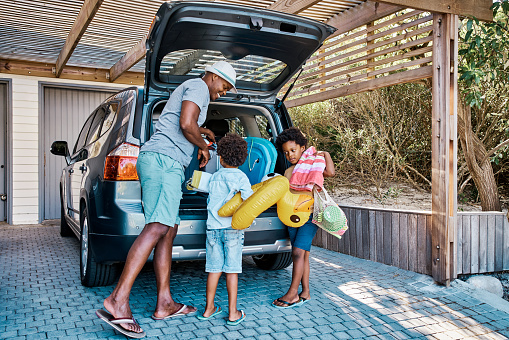Una familia empacando el coche para salir de vacaciones de verano. Un feliz padre afroamericano y sus dos lindos hijos pequeños preparando el equipaje en su vehículo para ir en un viaje por carretera, listos para el tiempo de viaje. photo