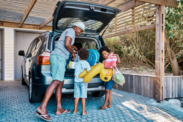 una familia empacando el coche para salir de vacaciones de verano. un feliz padre afroamericano y sus dos lindos hijos pequeños preparando el equipaje en su vehículo para ir en un viaje por carretera, listos para el tiempo de viaje. - road trip fotografías e imágenes de stock