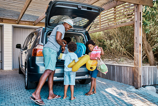 A family packing the car to leave for summer vacation. A happy african american father and his two cute little sons preparing the luggage in his vehicle to go on a road trip, ready for holiday time