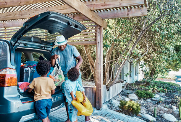 Excited black family packing their car trunk for a trip to the beach. Happy single dad and playful kids loading their boot for a summer vacation. Time to travel and have fun on a road trip together stock photo