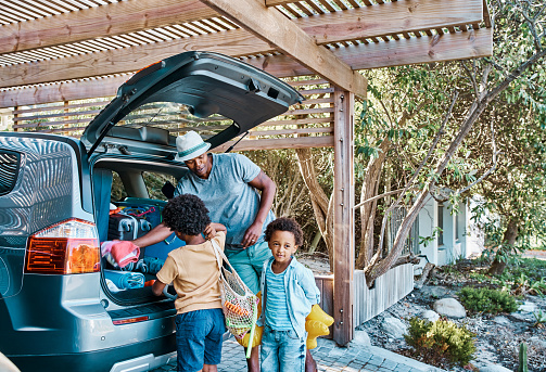 Family packing luggage in a car preparing to travel. Dad and little Afro American kids with baggage getting ready for the summer vacation. Father going on a road trip or to the beach with his son