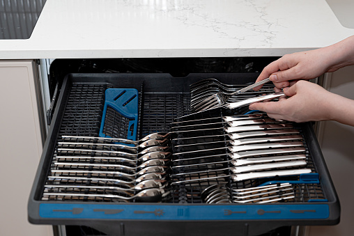 Person unloading open dishwasher machine with clean stainless cutlery set: knives, spoons and forks. Dishwashing machine with cutlery tray