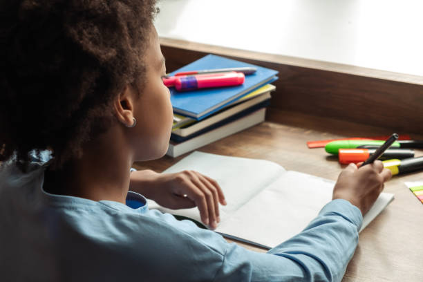 African-american preteen girl doing homework,making notes in notebook at home at her desk.Close up, rear view.Back to school concept.School distance education,home schooling,diverse people