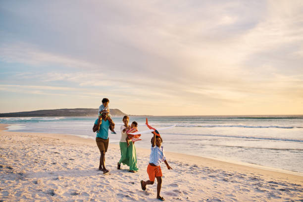 junge familie, die am wochenende zusammen am strand spazieren geht. liebevolle eltern bei einem spaziergang mit ihren kindern am meer während eines urlaubs. junge, der ein flugzeug fliegt und sich im urlaub mit mama und papa verbindet - family fun running couple stock-fotos und bilder