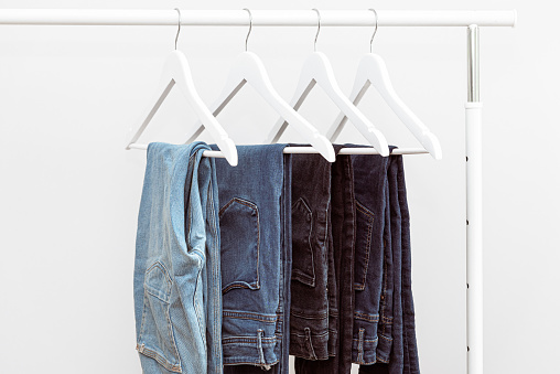 Many blue denim jeans hanging on white clothes hangers on clothing rack. Close up of folded casual denim jeans in wardrobe