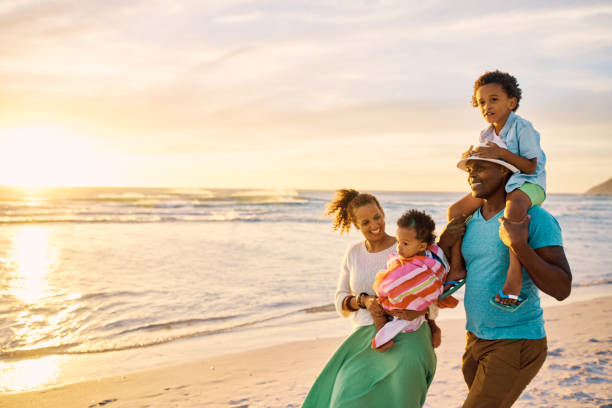 A black family enjoying their vacation, walking on the beach. Cheerful parents and their two kids traveling and enjoying a trip to the sea. Having fun together while on holiday in summer Happy black family walking on the beach and enjoying their vacation. Cheerful parents and their two kids strolling along the shoreline during sunset. Having fun together while on holiday in summer black couple sunset stock pictures, royalty-free photos & images
