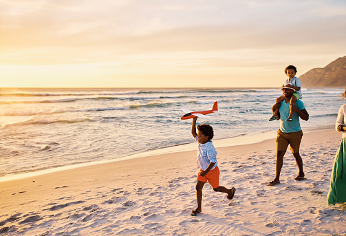 Carefree african american family walking and having fun together on the beach. Parents spend time with their kids while their son plays with his toy plane on holiday during a beautiful golden sunset