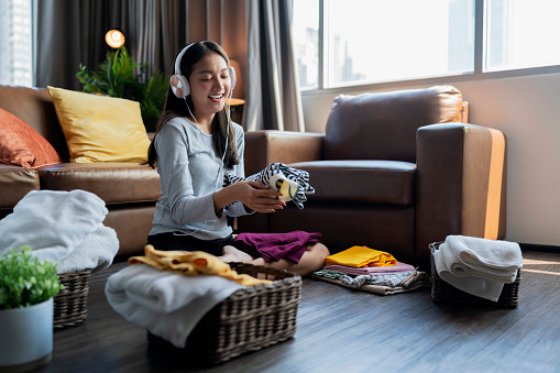Southeast Asian woman folding their cloth on weekend while listening music from headphone A young housewife woman touch a s tack of fresh washed clean linen with hands in living room at home