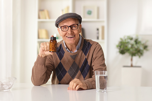 Happy elderly man sitting at home and showing a bottle of pills